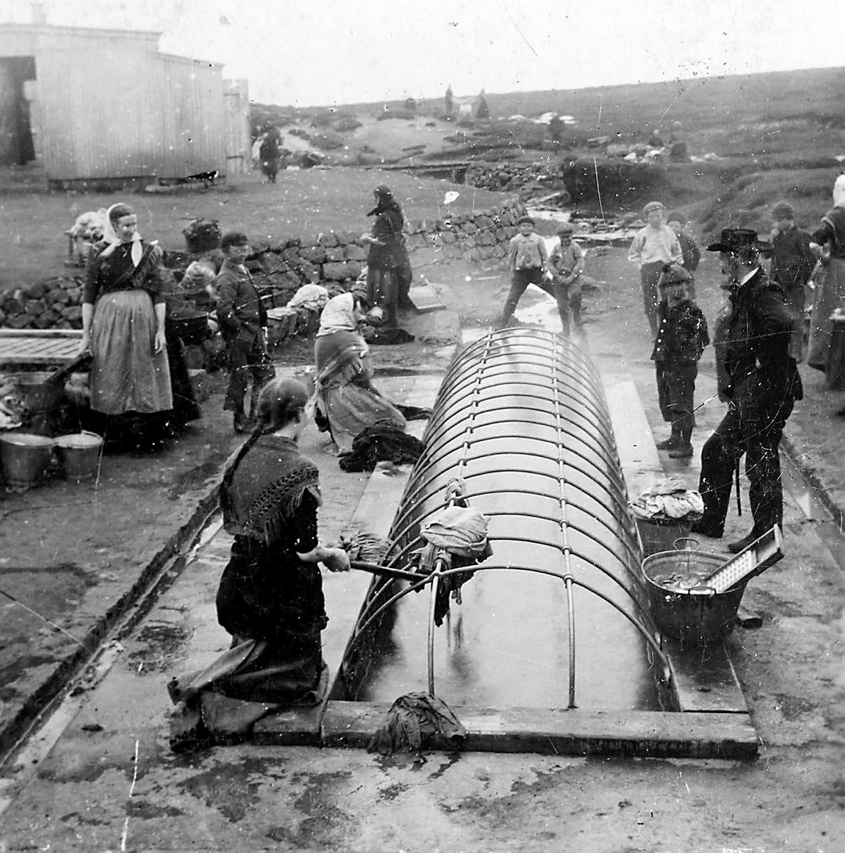 Konur þvo þvott í þvottalaugum í Laugardal. Karlar horfa á frá hægra megin. Svörthvít mynd frá aldamótunum 1900.